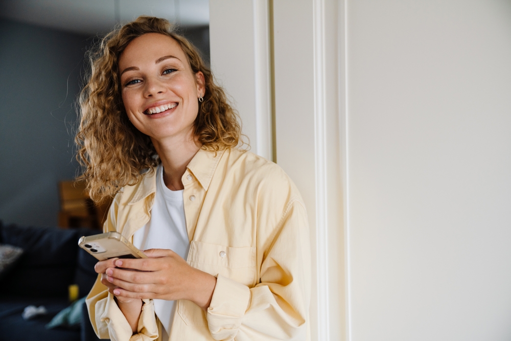 A woman with a bright smile holds a phone while standing indoors, showcasing her healthy teeth and radiant dental care - Dental Lab in Atlanta A woman with a bright smile holds a phone while standing indoors, showcasing her healthy teeth and radiant dental care - Dental Lab in Atlanta